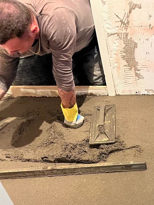 Sicil strijkt met gele handschoenen de nieuwe dekvloer van een badkamer af; achterin de douchegoot