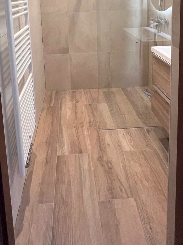 Na: dezelfde badkamer vanaf de deur, met houtlook vloertegels tot in de inloopdouche, een handdoekradiator en het wastafelmeubel