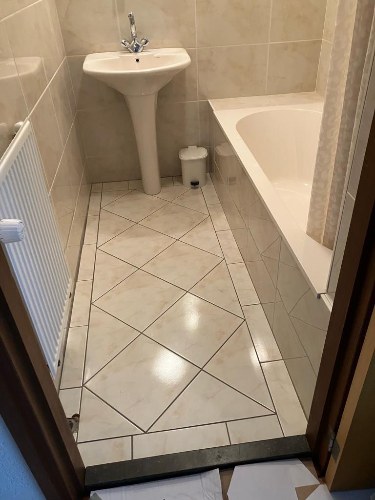 Voor: de oude badkamer vanaf de deur, met diagonaal gelegde beige vloertegels, een staande wastafel, een ligbad en een radiator